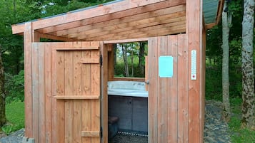 A wooden cabin with a door open, revealing a hot tub inside.