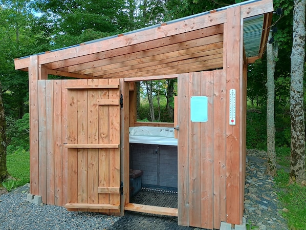 A wooden cabin with a door open, revealing a hot tub inside.