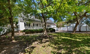 A white house with a porch, surrounded by trees and a fence. - Hillside by the Sea - 5BR, sleeps 14, game room (North Myrtle Beach)
