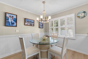 A dining area with a glass table surrounded by wicker chairs, a chandelier, and framed photographs on the wall. - Hillside by the Sea - 5BR, sleeps 14, game room (North Myrtle Beach)