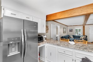 A kitchen with a stainless steel refrigerator, a microwave, and a water dispenser. There is a wooden ceiling and a living area with a sofa and a coffee table. - Hillside by the Sea - 5BR, sleeps 14, game room (North Myrtle Beach)