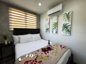 A bedroom with a bed, a window with blinds, an air conditioner, and wall art.