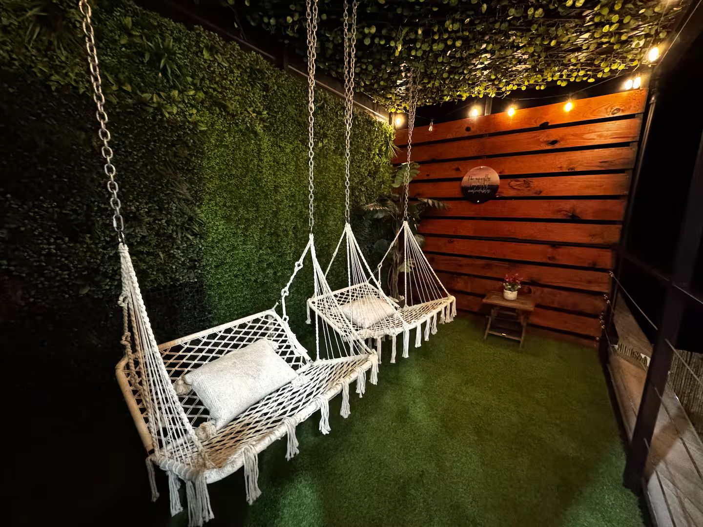 Une pièce avec deux hamacs en macramé blanc, une petite table en bois ornée d’une plante et un mur vert agrémenté de guirlandes lumineuses.