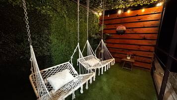 Une pièce avec deux hamacs en macramé blanc, une petite table en bois ornée d’une plante et un mur vert agrémenté de guirlandes lumineuses.