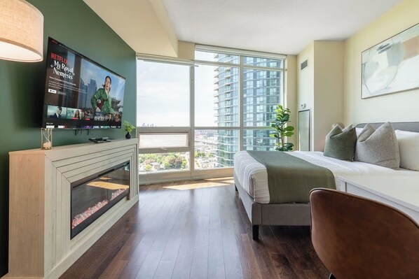 A modern hotel room with a large bed, a television, and a view of high-rise buildings. - Sleek Studio in Fort York Near Lake & Liberty (Toronto)