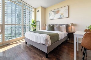 A modern bedroom with a large window, a bed with multiple pillows, a bedside table with a lamp, and a painting on the wall. - Sleek Studio in Fort York Near Lake & Liberty (Toronto)