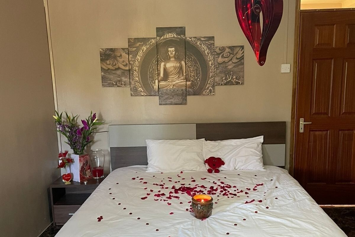 Quarto de hotel com uma cama, decoração na parede, luminária em formato de coração e mesa de cabeceira com um vaso de flores.