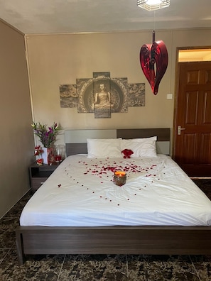 Quarto de hotel com uma cama, decoração na parede, luminária em formato de coração e mesa de cabeceira com um vaso de flores.