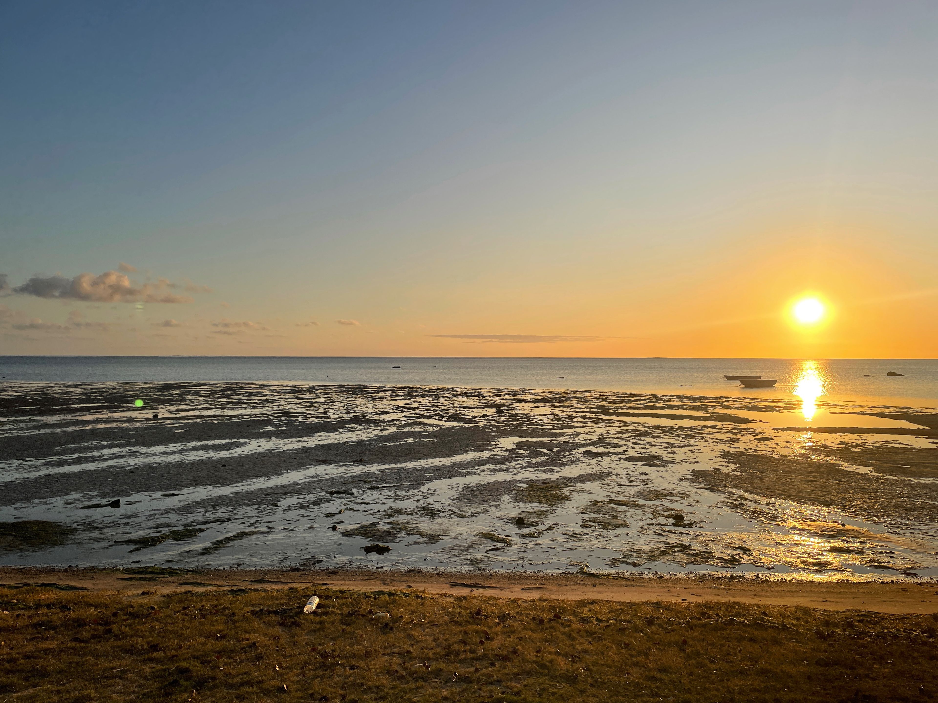 A beach with a clear sky, the sun setting over the horizon, and boats on the water.