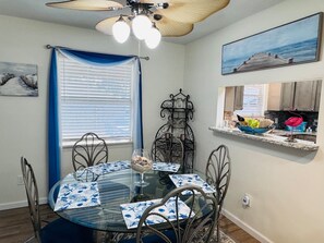 A dining area with a glass table, metal chairs, a ceiling fan, and a kitchen counter with a window view. - Coastal Hangout | Hot Tub | GameRm | Mins2Bch (Panama City)