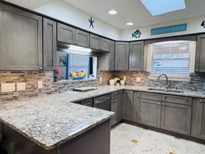 A kitchen with granite countertops, dark cabinetry, a window with blinds, and a decorative sign above the window. - Coastal Hangout | Hot Tub | GameRm | Mins2Bch (Panama City)