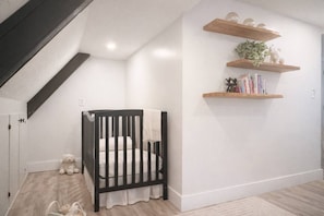 A room with a black crib, wooden shelves, and a white wall. - One-Block Beach Oasis w/ Poolside Game & Golf Cart (Panama City Beach)