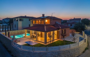 Una casa de dos pisos con piscina, un patio cubierto y un muro de piedra que rodea la propiedad.