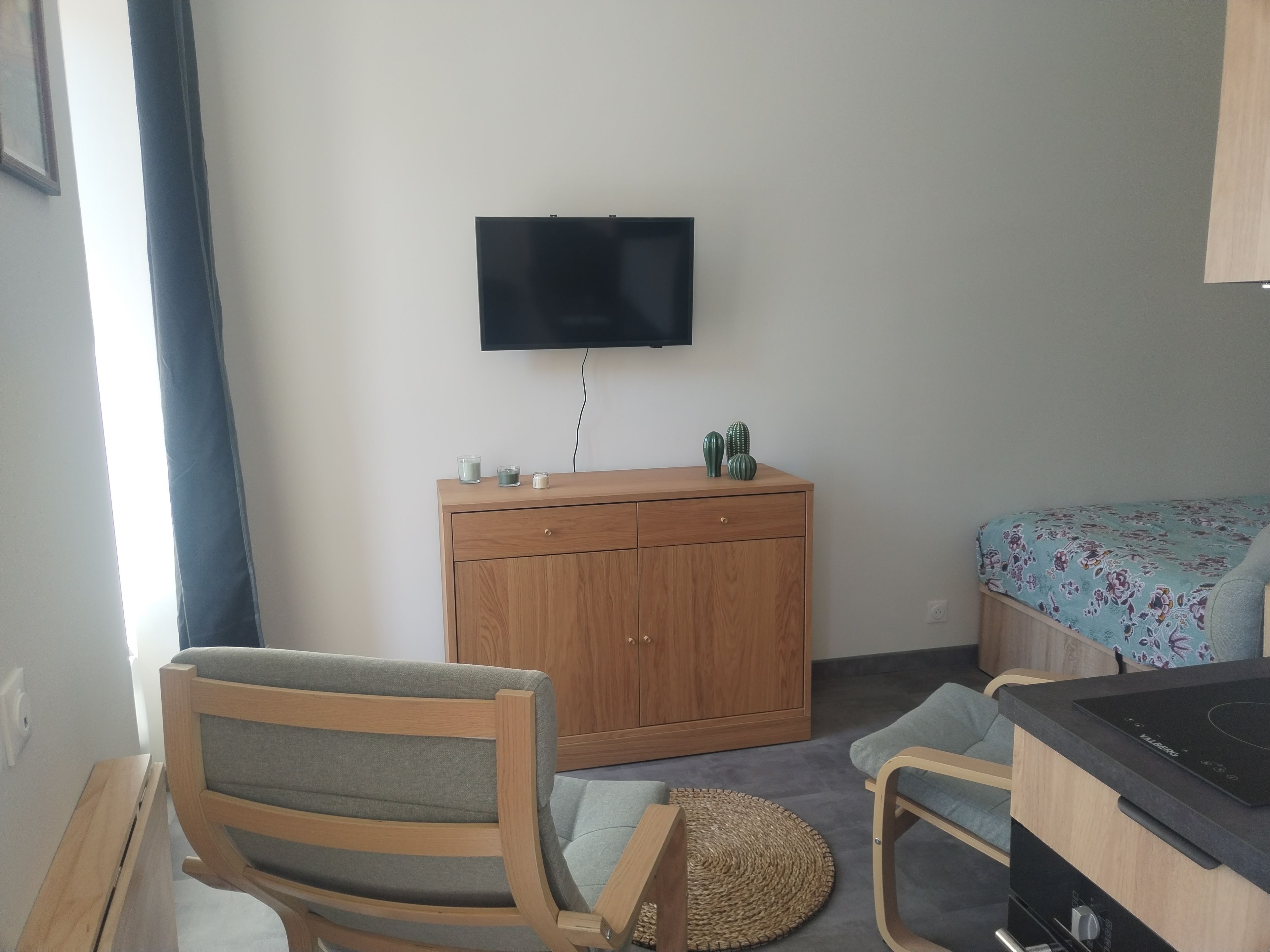 Ein Zimmer mit einem Holzkasten, einem an der Wand montierten Fernseher, einem Bett mit einer blumigen Tagesdecke und einem Sessel mit einem gewebten Sitz.