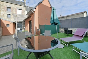 Ein Garten mit einem Glastisch, zwei Liegestühlen, einem grünen Sonnenschirm und einem Steinhaus.