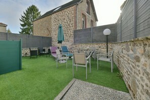 A patio area with artificial grass, outdoor furniture, and a stone building in the background. - Le Lilas - Petfriendly - Jacuzzi, sauna (La Ferrière-aux-Étangs)