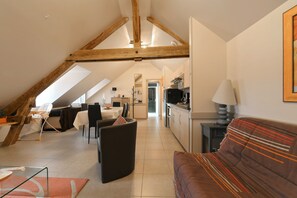 Een ruime loft met een hoge plafond, zichtbare houten balken, een eethoek, een keuken en een zithoek met een bank.
