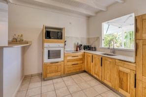 A modern kitchen with wooden cabinets, a built-in microwave, and a window with a view of greenery. - Villa Rosalie - Piscine - Proche plage (Le Diamant)