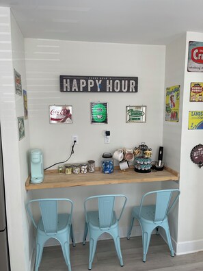 A kitchen area with a coffee maker, jars, and a 'Happy Hour' sign. There are three chairs and various vintage posters on the wall. - Admiral's Suite 300ft to Beach and Boardwalk (Seaside Heights)
