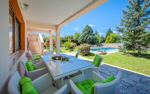 Um pátio externo com mesa de vidro e cadeiras de vime, piscina e um muro de pedra ao fundo.