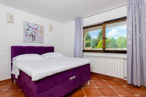 Quarto com cama roxa, roupa de cama branca, janela com cortinas, radiador e um quadro com motivos florais na parede.