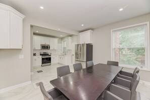 Una cocina moderna con armarios blancos, electrodomésticos de acero inoxidable y un comedor con mesa de madera oscura y sillas grises.