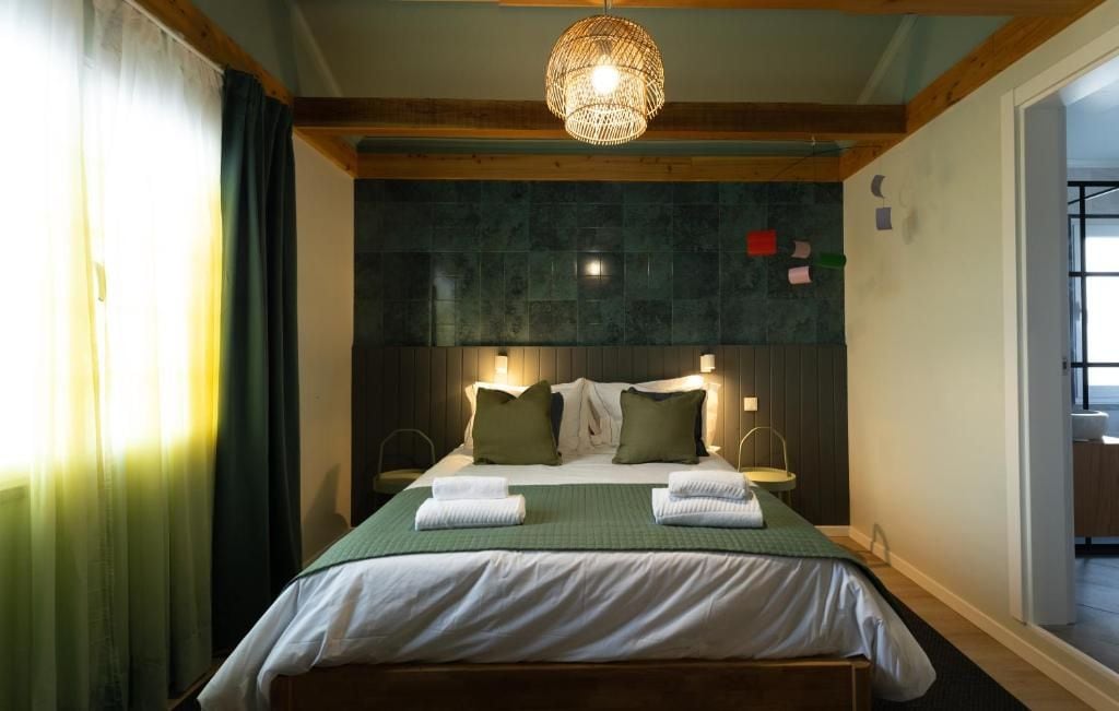 Quarto de hotel com cama, luminária pendente, janela com cortinas e porta que dá para outro cômodo.
