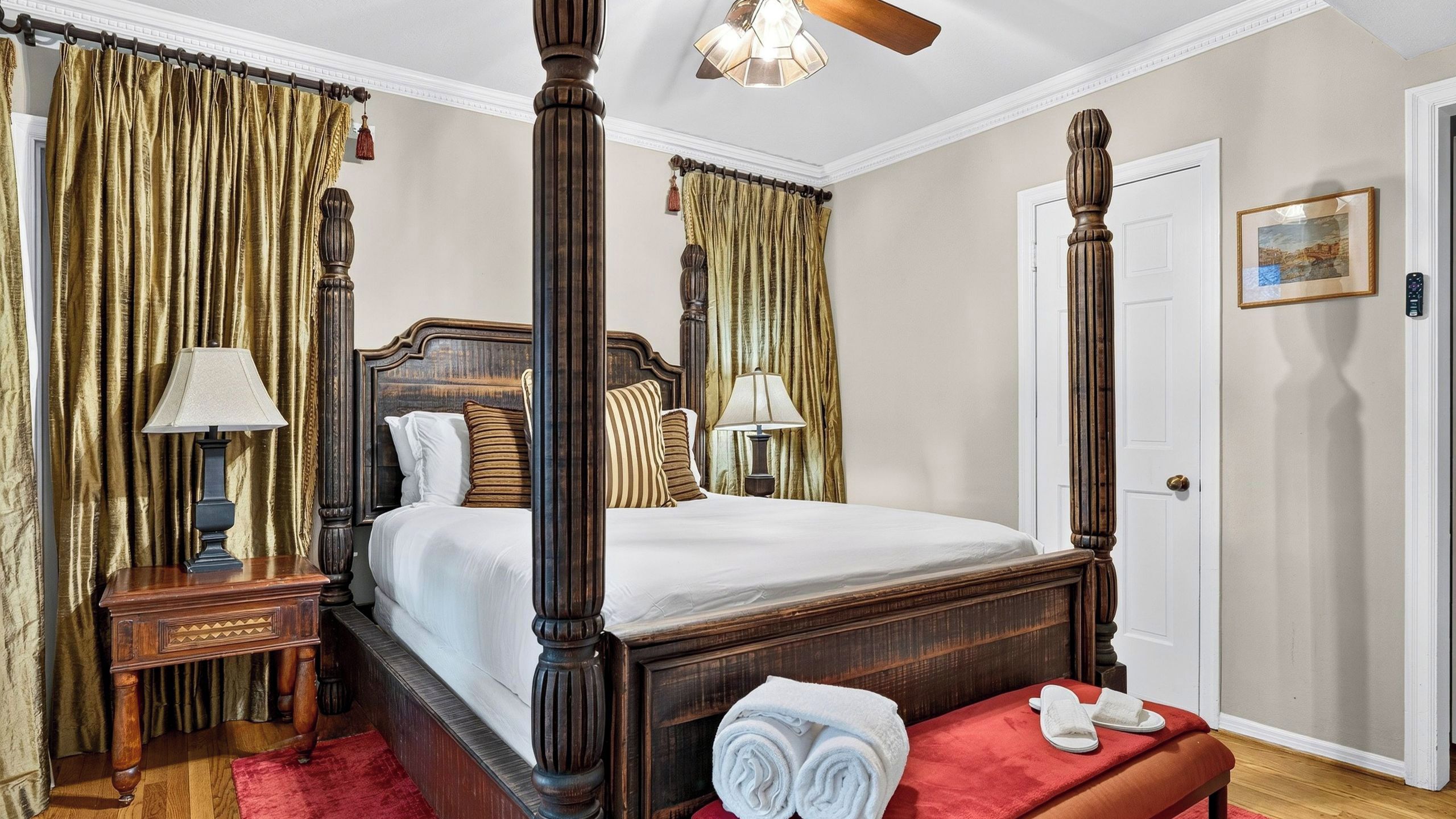 A four-poster bed with a wooden headboard, two bedside tables with lamps, a red bench with towels, and a door with a picture frame.