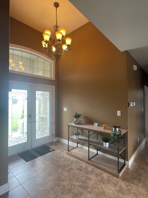 A hallway with a chandelier, a glass door entrance, a console table with plants, and a tiled floor.