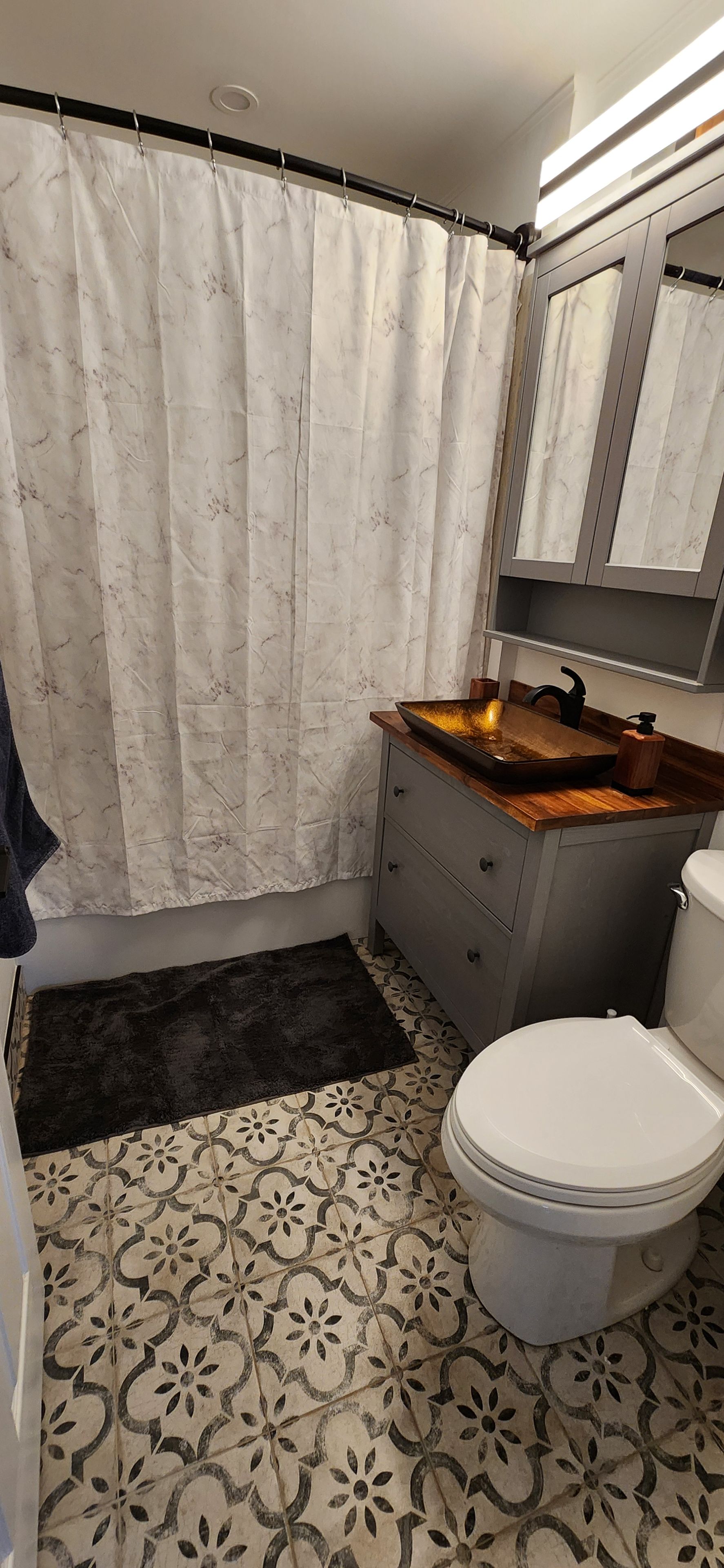 A bathroom with a shower curtain, a sink with a wooden countertop, a toilet, and patterned floor tiles.