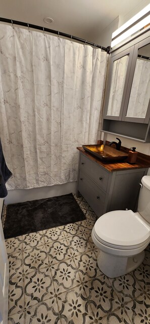 Ein Badezimmer mit Duschvorhang, Waschbecken mit Holzauszug, Toilette und fugenlosen Bodenfliesen.