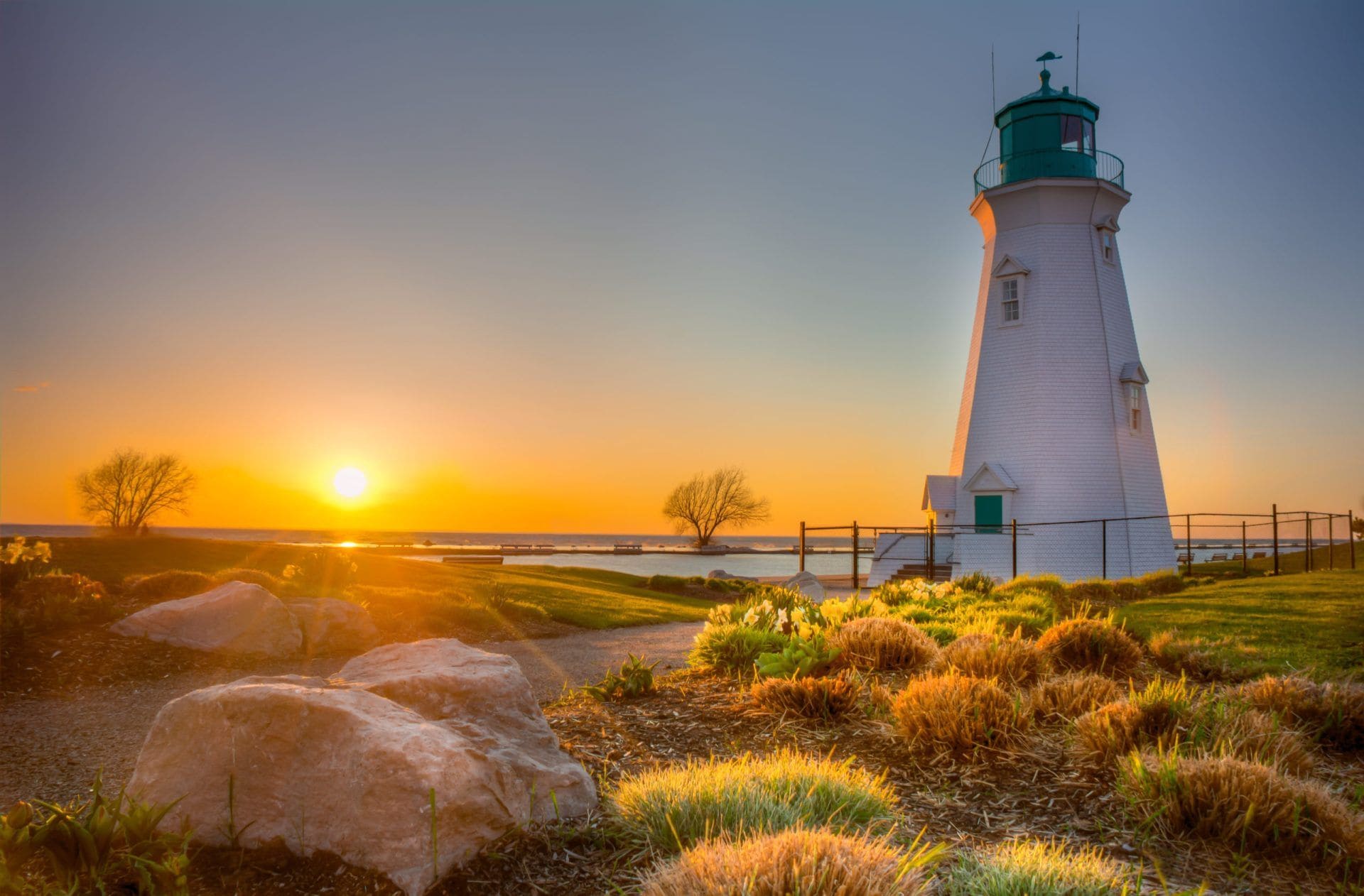 A lighthouse with a green top, surrounded by rocks and grass, with the sun setting in the background.