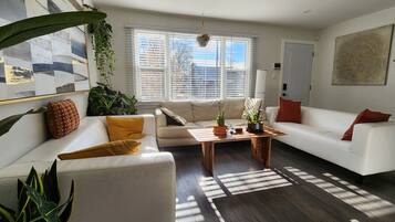 A modern living room with a white sofa, a wooden coffee table, and a large window with blinds.