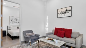 A modern living room with a bed, a grey sofa with red cushions, a coffee table with food, and artwork on the wall. - Exeter House by Avantstay Sleeps 18 + Casita Pool + Spa + Theater Camelback Views (Phoenix)