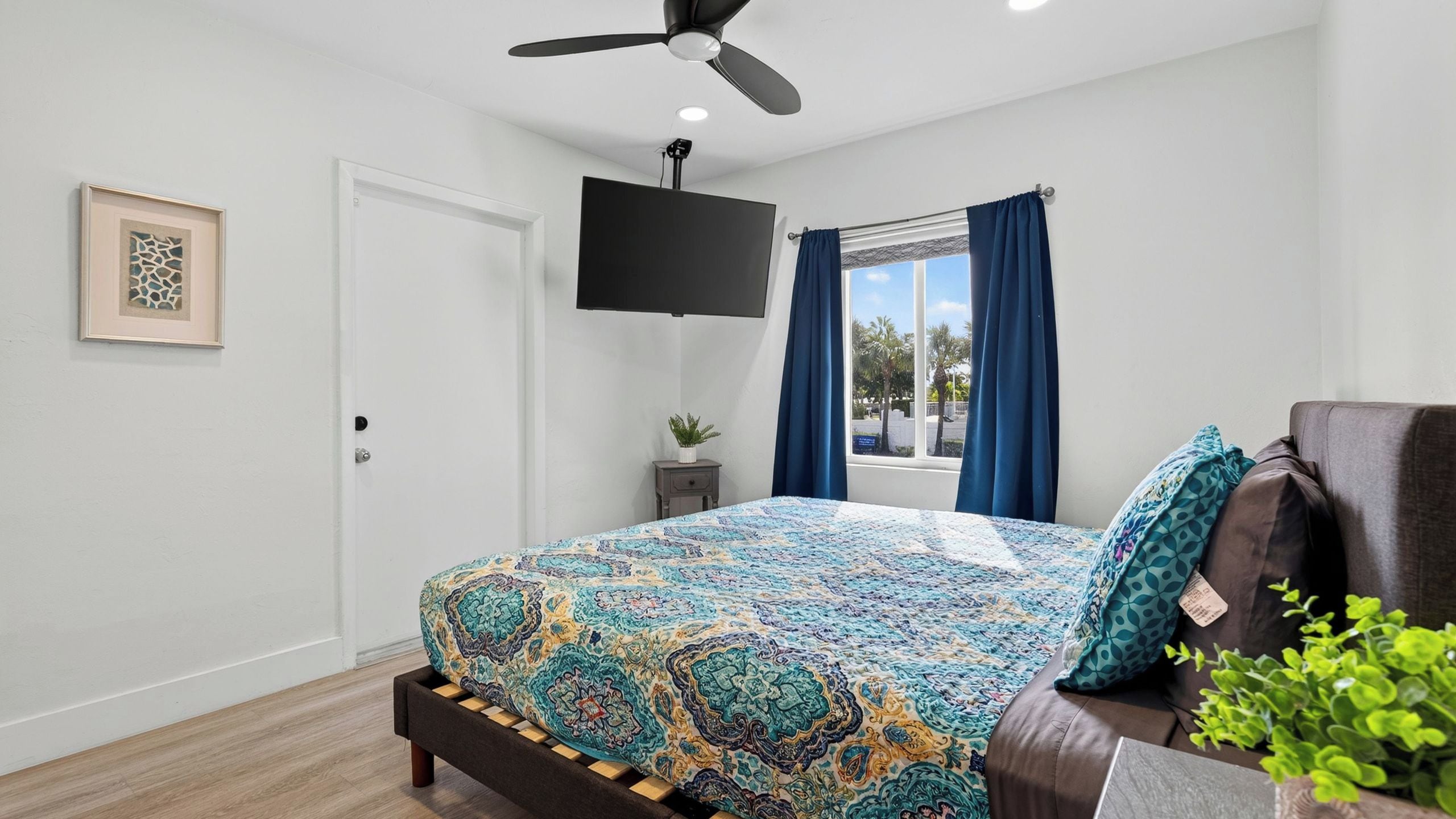 A bedroom with a bed, a TV mounted on the wall, a window with curtains, and a ceiling fan.