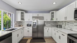 A modern kitchen with white cabinets, a stainless steel refrigerator, and a window with a view of greenery. - Lauderdale Oasis by Avantstay 3-min Walk To Beach Hot Tub + Private Yard Near Las Olas (Fort Lauderdale)