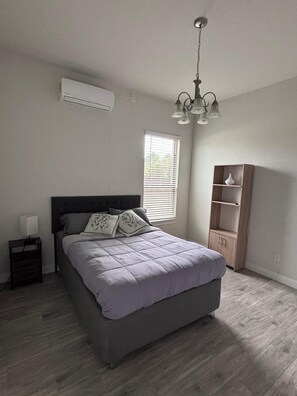 Een slaapkamer met een bed, een nachtkastje, een raam met jaloezieën, een ingebouwde houten plank en een plafondlamp.