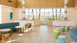 A modern lobby with a blue sofa, wooden chairs, a marble reception desk, and a large window with a view. - Spacious 2BR Villa at Myrtle Beach Oceanfront – Full Kitchen & Resort Fun (Myrtle Beach)