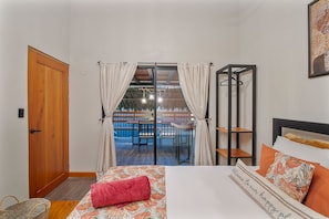 Ein Schlafzimmer mit einem Bett, einer Holztür, einem Fenster mit Vorhängen, einem Kleiderständer und Blick ins Freie.