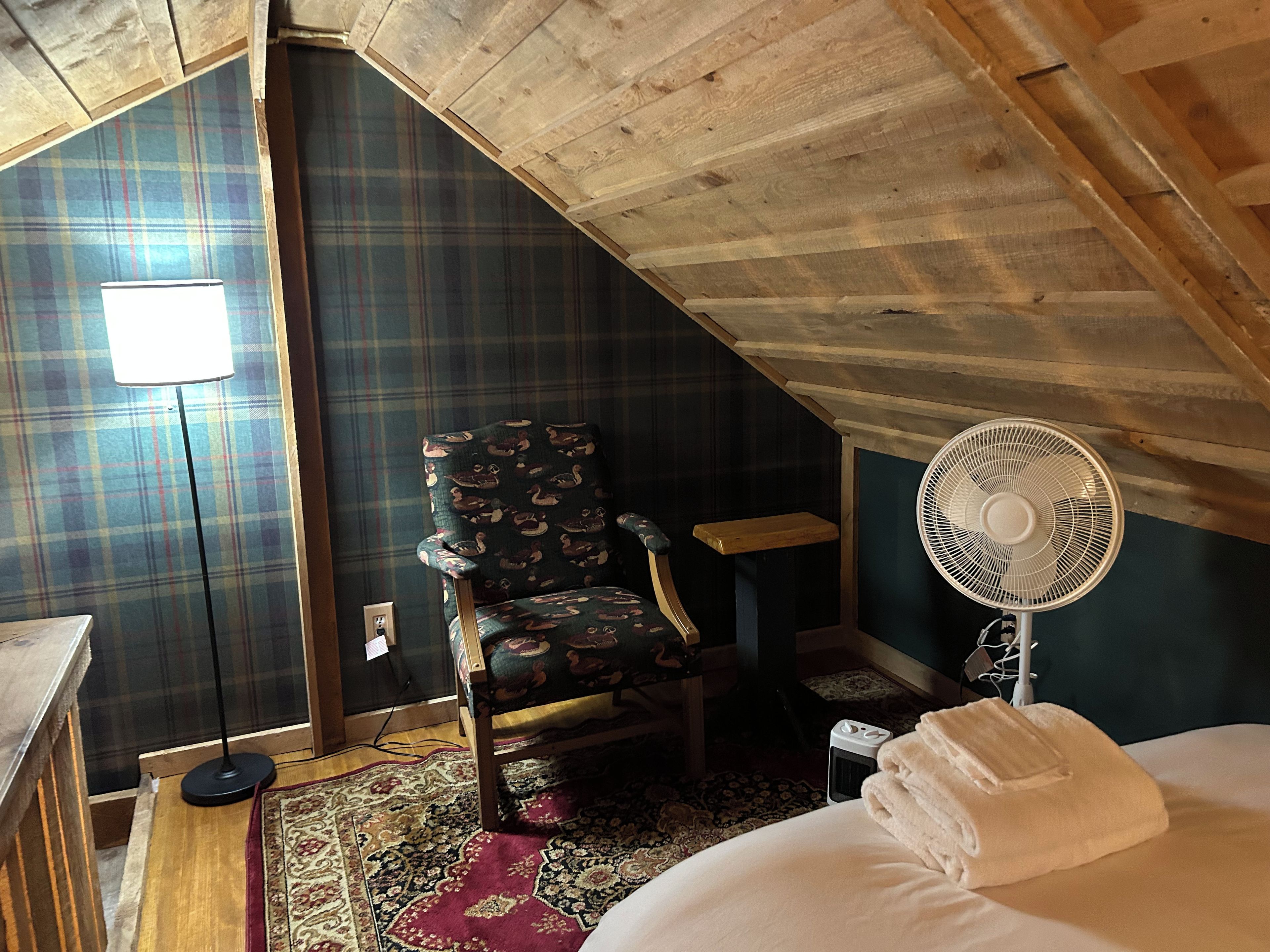 A room with a sloped ceiling, a plaid-patterned wall, a wooden chair, a standing fan, a bed with a white blanket, and a small table.