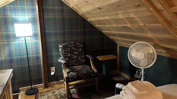 A room with a sloped ceiling, a plaid-patterned wall, a wooden chair, a standing fan, a bed with a white blanket, and a small table.