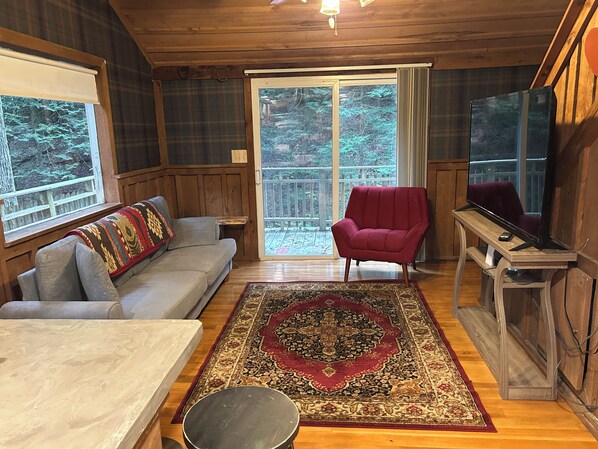 A cozy living room with a sofa, a red armchair, a television, and a large window with a view of a wooden deck.