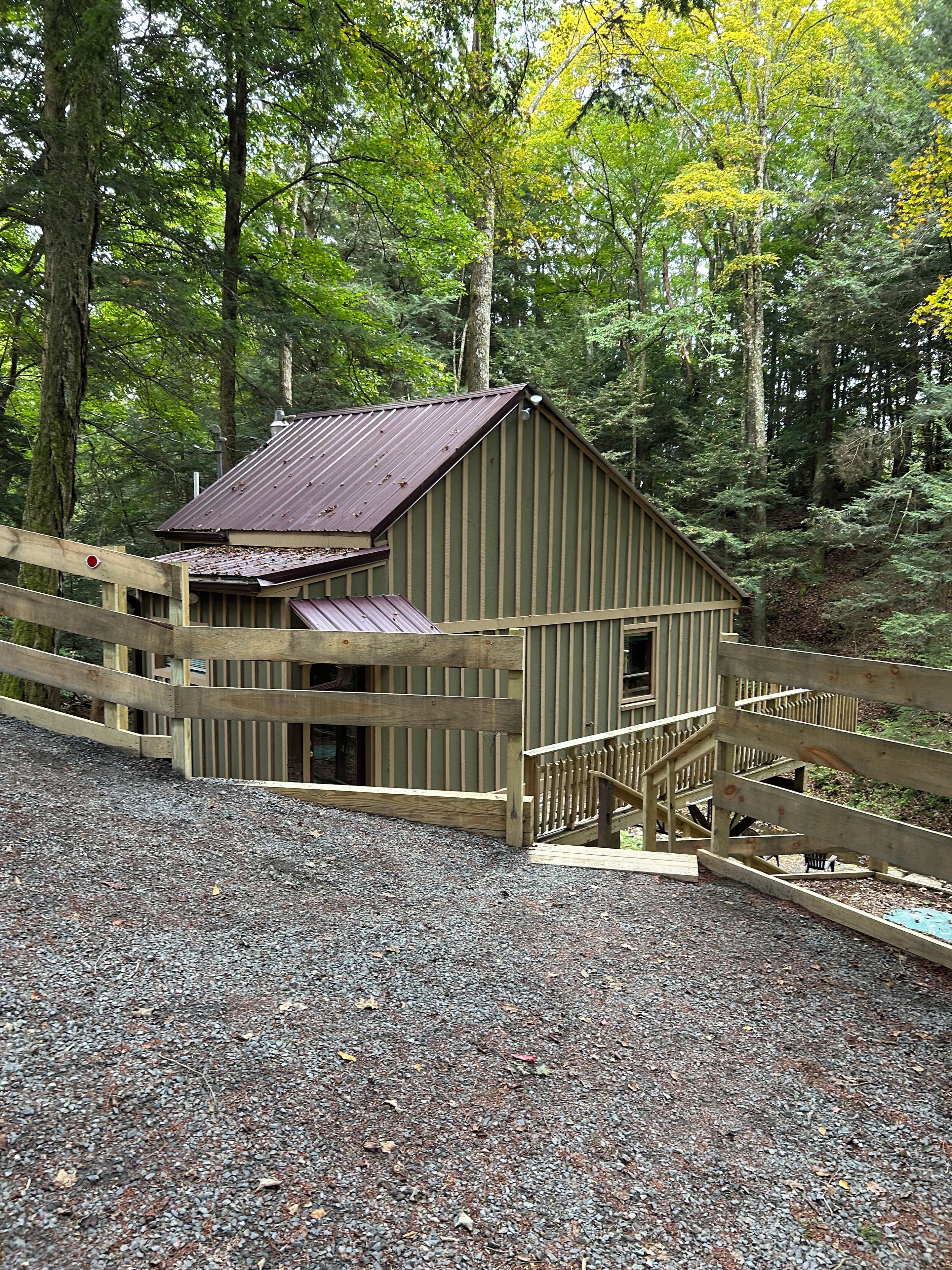A wooden cabin with a metal roof, surrounded by a wooden fence, set in a forested area with gravel ground.