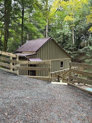 A wooden cabin with a metal roof, surrounded by a wooden fence, set in a forested area with gravel ground.
