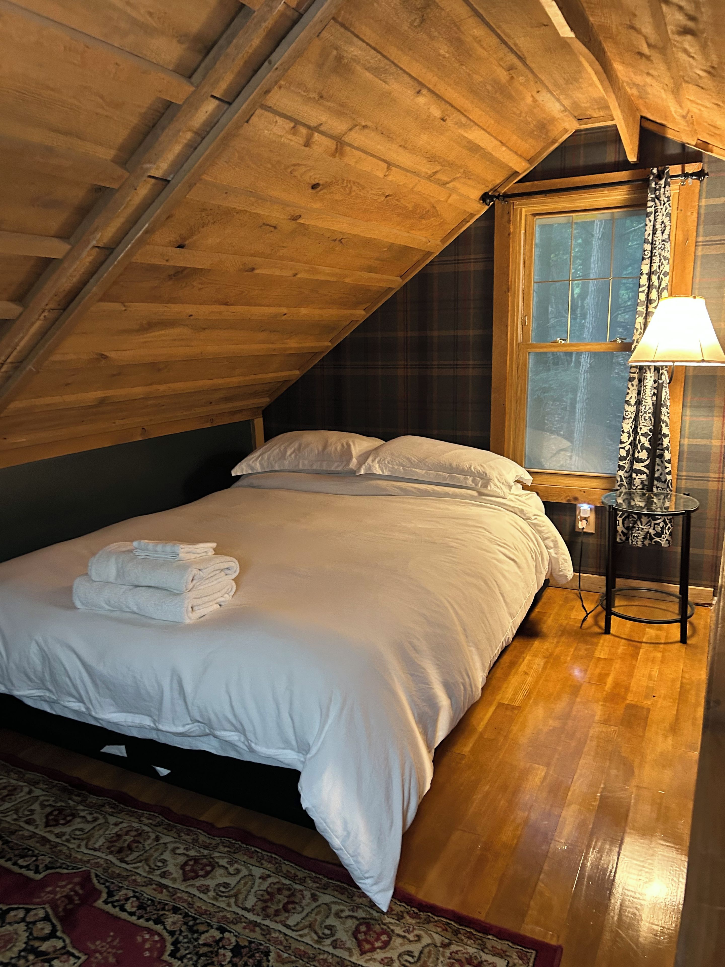 A bedroom with a wooden ceiling, a bed with white linens, a window with curtains, and a bedside lamp.