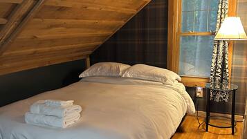 A bedroom with a wooden ceiling, a bed with white linens, a window with curtains, and a bedside lamp.