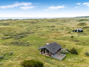 Eenlaags huis met een donker dak, omgeven door een grasveld met glooiende heuvels op de achtergrond.