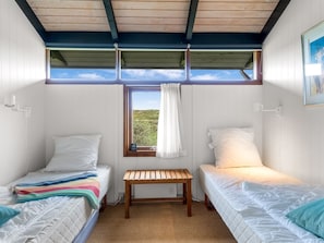 Quarto com duas camas de solteiro, um banco de madeira, janela com cortinas e vista para o verde lá fora.