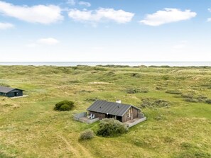 Uma casa de madeira térrea, com telhado plano, cercada por dunas gramadas e um céu claro.