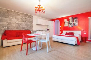 A modern living room with a red sofa, a white dining table with chairs, a kitchen area, and a red accent wall with artwork. - Kedimore - Red Apartment (Verona)